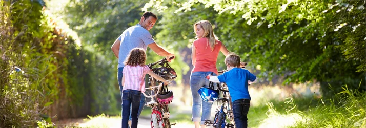 People viewed from behind walking bicycles on a grassy path, holding helmets, with trees in the background.