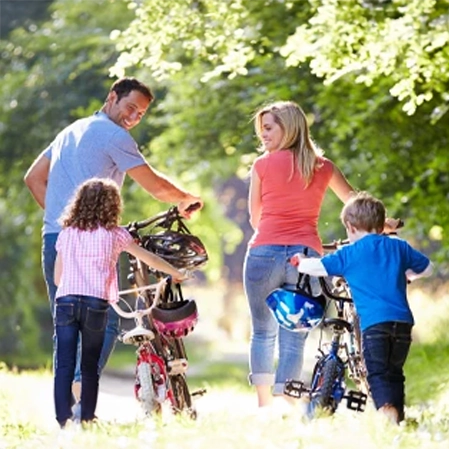 Two adults and two children walking bicycles along a sunlit path surrounded by trees.
