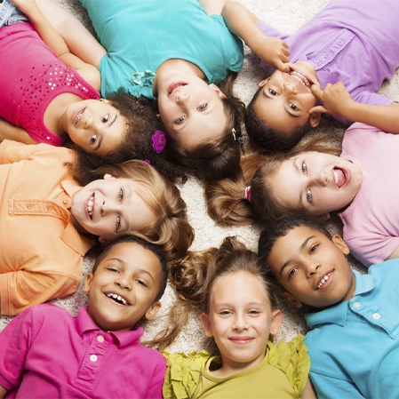 Group of children lying on a carpet in a circle, heads together, wearing brightly colored shirts.