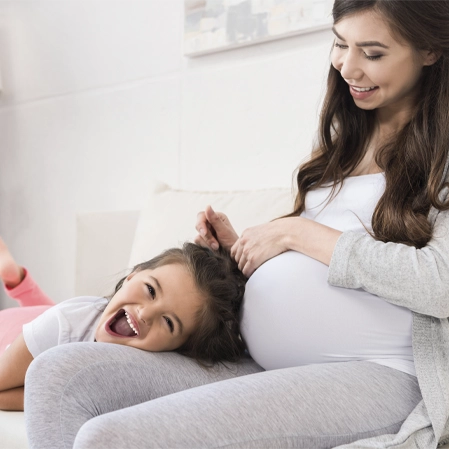 Pregnant person seated on a sofa while a child rests beside the belly, with hands arranging the child's hair.